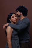 A man and a woman in a studio setting against a solid dark brown background. The woman wears a sleeveless dark gray dress, and the man wears a dark gray long-sleeve knit sweater and brown pants.