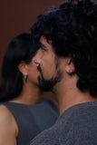 A close-up side profile of a man with curly black hair and a beard, wearing a gray knit shirt, with a woman in a dark gray sleeveless top blurred in the background against a warm brown wall.