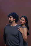 A man and a woman posing against a solid dark reddish-brown background. The man in the foreground wears a dark gray long-sleeve crewneck sweater, while the woman behind him wears a dark gray sleeveless top.