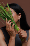 A woman with long black hair holds a bunch of green grass near her face, wearing a dark gray sleeveless top and a dangling silver earring, against a solid brown background.