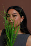 A woman with long dark hair wearing a dark gray sleeveless top and a silver hoop earring, holding green ornamental grass in front of her face against a solid brown background.