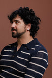 A man with curly black hair and a beard wearing a navy blue and white striped knit polo shirt, photographed against a solid warm brown background.