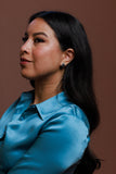 A woman with long dark hair wearing a shiny light blue satin button-up shirt, shown in profile against a solid brown background.