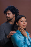 A man with curly dark hair and a beard wearing a gray long-sleeve shirt stands behind a woman with dark hair wearing a shiny light blue satin top, posing with her hand on his shoulder against a solid brown studio background.