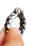 A close-up of a silver-toned twisted earring held by a person's fingers, showing a circular brand stamp.