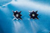 A pair of star-shaped stud earrings with a polished silver-tone finish and a small clear crystal in the center, displayed on a textured blue surface.