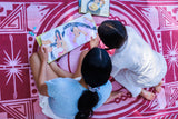 Two children sitting on a pink patterned blanket, reading a colorful illustrated book together from an overhead view.