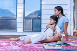 A woman and a young child sit together on a pink patterned blanket in front of a white house with large windows. The woman wears a light blue t-shirt and white shorts, and the girl wears a white t-shirt and white pants.