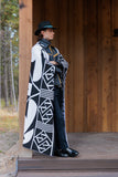 A person wearing a long black-and-white geometric patterned cape or wrap, dark pants, black boots, and a black hat with a turquoise band, standing on a wooden porch in front of a wood-paneled building.
