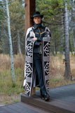 A man wearing a long black and white geometric patterned coat, a black hat, and a dark shirt stands on a wooden deck in an outdoor wooded setting.