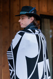 A man wearing a black wide-brim hat with a blue feather and a black-and-white geometric patterned blanket draped over his shoulders, standing in front of a wooden wall.