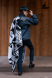 A man stands on wooden steps wearing a black hat with a turquoise band, a dark denim jacket with gold lettering on the back, dark trousers, and black boots. A large black-and-white geometric patterned blanket is draped over his shoulders.