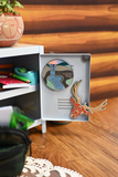 A miniature white school locker decoration with its door open, featuring a circular magnet and a red-orange elk head ornament attached to the door, set on a wooden floor next to a small shelf.