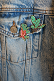 A close-up of a denim jacket showing a decorative enamel pin shaped like a red bleeding heart with green leaves, pinned to the front pocket.