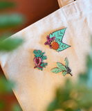 Three colorful embroidered botanical-themed enamel pins displayed on a beige canvas tote bag, with soft green leaves and a warm brown background blurred in the foreground.