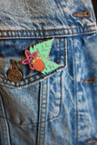A close-up of a denim jacket pocket showing a colorful enamel lapel pin shaped like a stylized berry with pink and green details, next to a bronze-tone button.