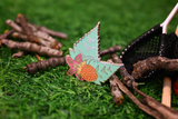 A colorful enamel pin with a mint green background, pink and orange design, and white geometric patterns, resting on green grass among twigs and a black net.