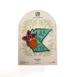 An enamel pin featuring a stylized salmon berry with a green leaf and purple flower, mounted on a branded display card from Eighth Generation.