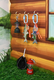 A set of fishing-themed keychains hanging from gold hooks on a wooden wall, displayed alongside a small red lantern and a black net on artificial grass.