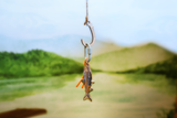 A small keychain shaped like a fish hanging from a wooden hook and clip against a blurred outdoor background of green hills and a pale sky.