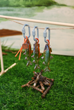 Three small fish-shaped keychains with gray clips hang from a string over a miniature campfire made of twigs, set on artificial green grass with a blurred outdoor-style background.