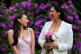 Two women standing in front of purple flowers, one holding flowers.
