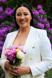Woman in a white blazer holding flowers with purple flowers in the background