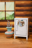 A miniature white metal locker toy or collectible display piece with a circular magnet, placed on a wooden surface next to a small blue and orange patterned vase with dried stems. The background shows a rustic wood-paneled wall and window.
